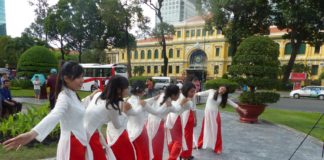 Das Postamt in Saigon aus dem 19. Jahrhundert: Es war ein Architekt daran beteiligt, den Sie alle kennen. – ‚Reihe Postämter der Welt‘, Nr.2 Vietnamesinnen einer Tanzgruppe mit roten Hosen, leichten, weißen Übergewändern und schwarzen Schuhen tanzen auf einem glatten Weg aus verschiedenfarbigen, quadratischen, marmorierten Natursteinfliesen in der gepflegten Grünfläche oder einem kleinen Park mit rundgeschnittenen, dunkelgrünen Bäumchen, z.T. in großen, auf dem kreisrunden Weg stehenden Tonschalen in Richtung eines Zebrastreifens, der über eine Straße, auf der Busse fahren, zum Postamt Saigons führt, einem gelben, zweistöckigen, repräsentativen, französischen Altbau mit einem dreietagigen Mittelbau. - Symbolisch: Müssen wir jetzt nach Saigon zur Post, weil die deutschen Postämter geschlossen werden? Wie weit?