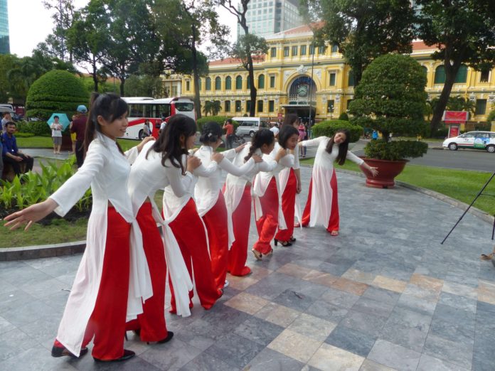 Vietnamesinnen einer Tanzgruppe mit roten Hosen, leichten, weißen Übergewändern und schwarzen Schuhen tanzen auf einem glatten Weg aus verschiedenfarbigen, quadratischen, marmorierten Natursteinfliesen in der gepflegten Grünfläche oder einem kleinen Park mit rundgeschnittenen, dunkelgrünen Bäumchen, z.T. in großen, auf dem kreisrunden Weg stehenden Tonschalen in Richtung eines Zebrastreifens, der über eine Straße, auf der Busse fahren, zum Postamt Saigons führt, einem gelben, zweistöckigen, repräsentativen, französischen Altbau mit einem dreietagigen Mittelbau. - Symbolisch: Müssen wir jetzt nach Saigon zur Post, weil die deutschen Postämter geschlossen werden? Wie weit?