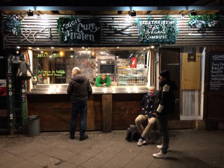 Der vegane Imbiss "Veggie CURRY-Piraten" am S-Bhf-Ausgang Frankfurter Allee. Man sieht den Imbiss frontal ganz, eine Angestellte blickt durchs Fenster, 3 Gäste stehen und sitzen vor dem Imbiss